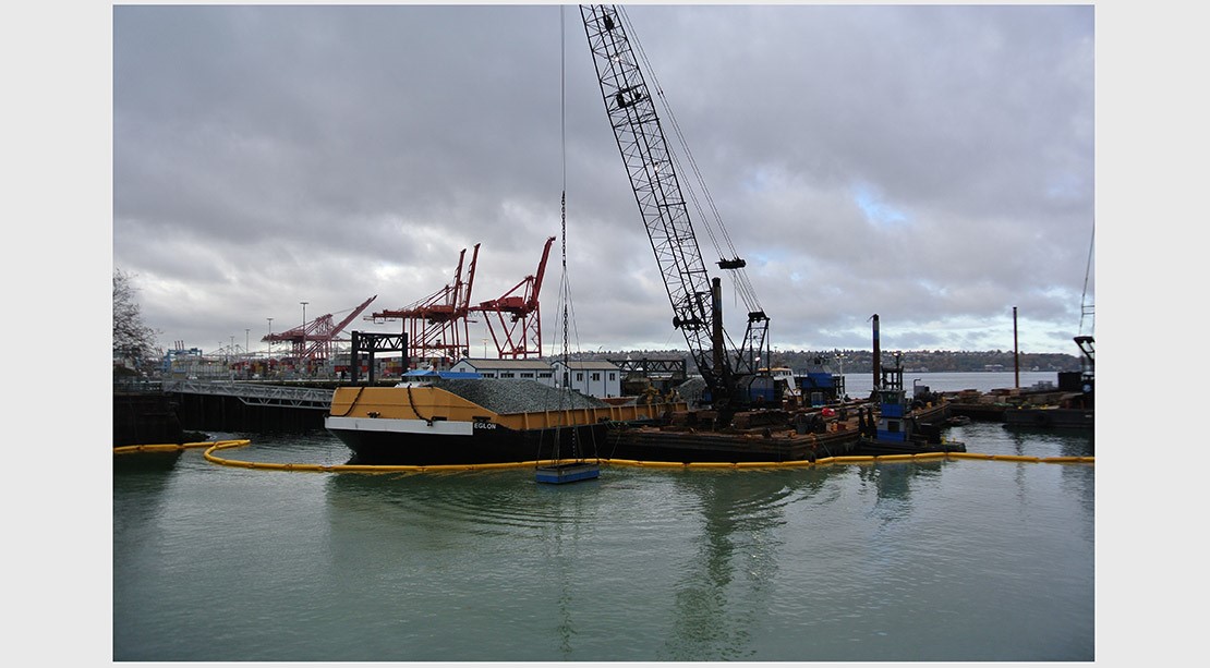 Photo of a crane on a barge lowering a pile of rocks into the marine habitat zone
