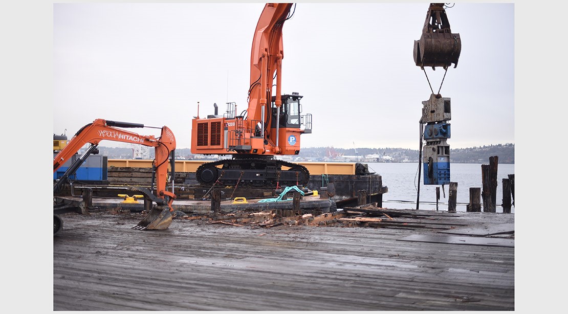 Photo of a square barge with a vibratory pile driver on-top installing new steel piles. 