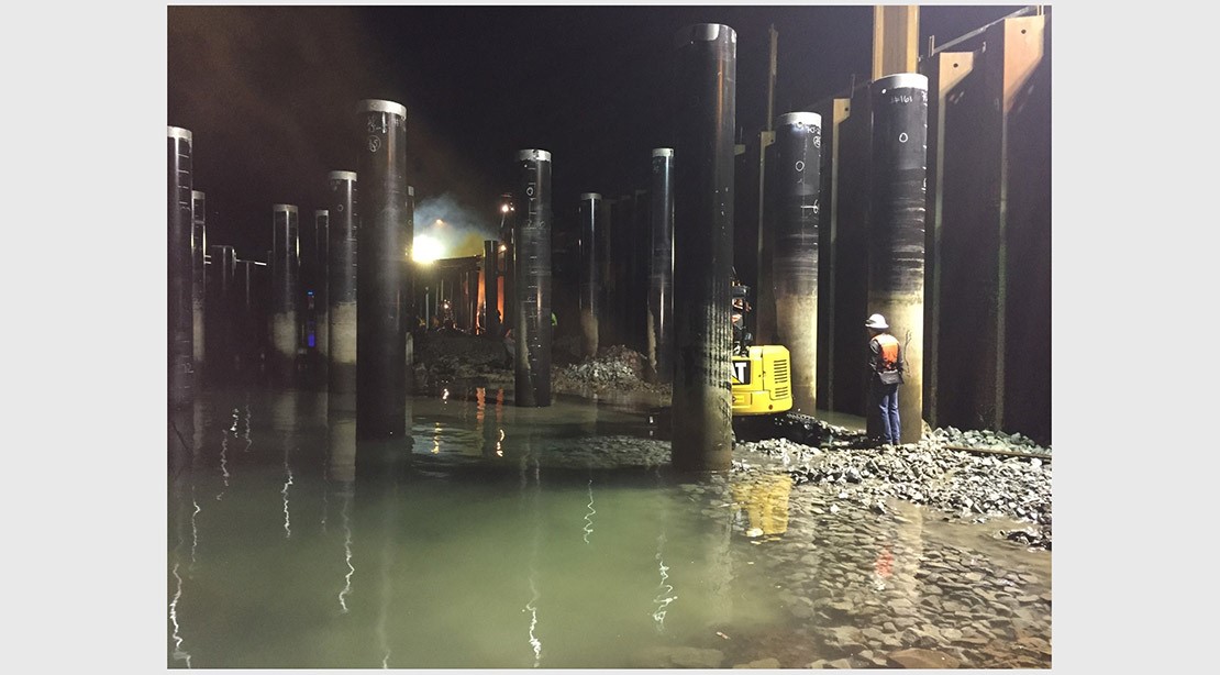 Photo of a crew member standing beside the new steel piles at low tide at night. 