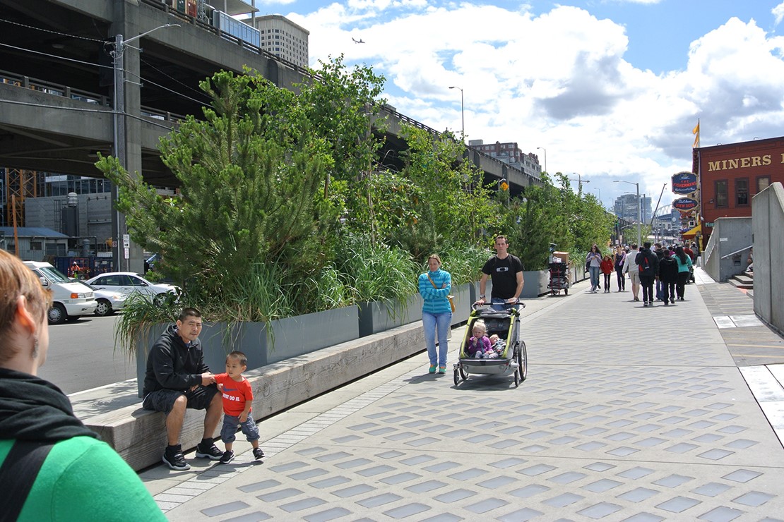 Photo shows people walking on a sidewalk with a light penetrating surface, including a family with a stroller next to green leafy plants.  