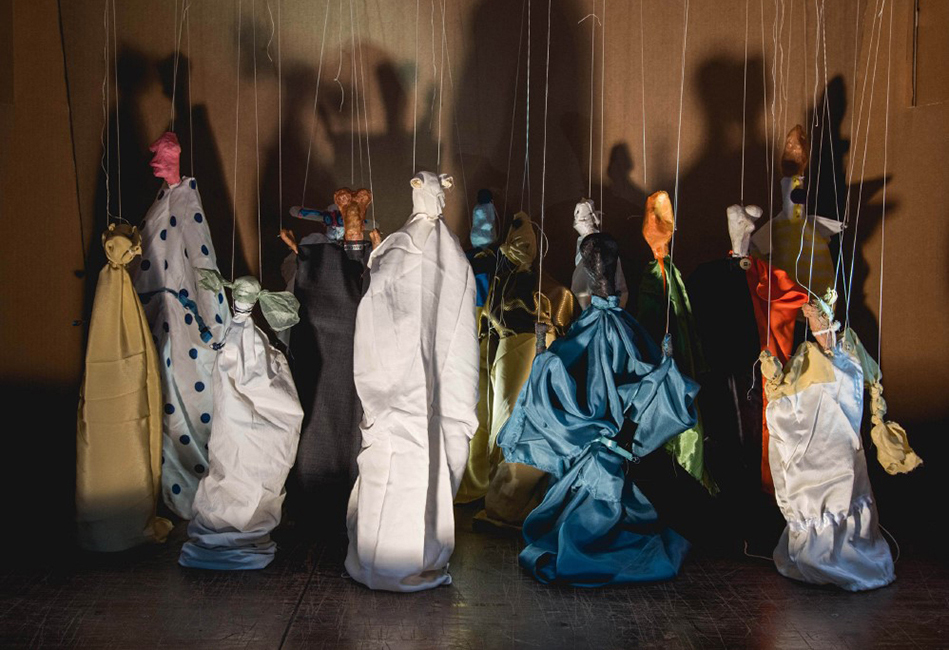 Photo of a small scale replica of puppet cloth figures being held up by strings. The puppets are of all different colors and are lit up, which creates larger shadows on the wall behind them.