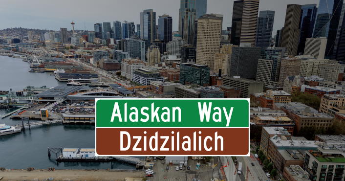 Road sign reading Alaskan Way on top and Dzidilalich on bottom superimposed over an aerial view of the Seattle Waterfront