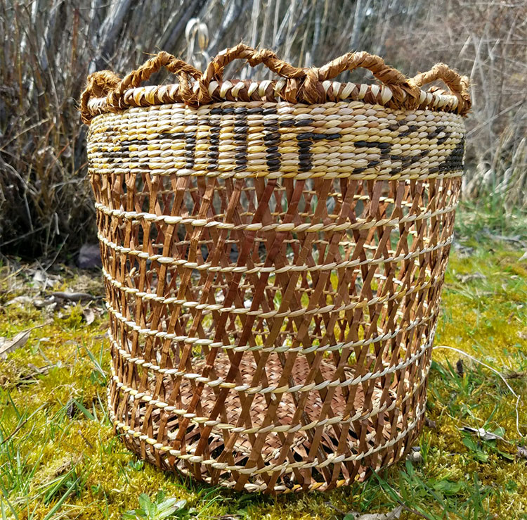 A close-up of a brown basket sitting on grass with large see-through weaving. It has tighter weaving at the top with black symbols.